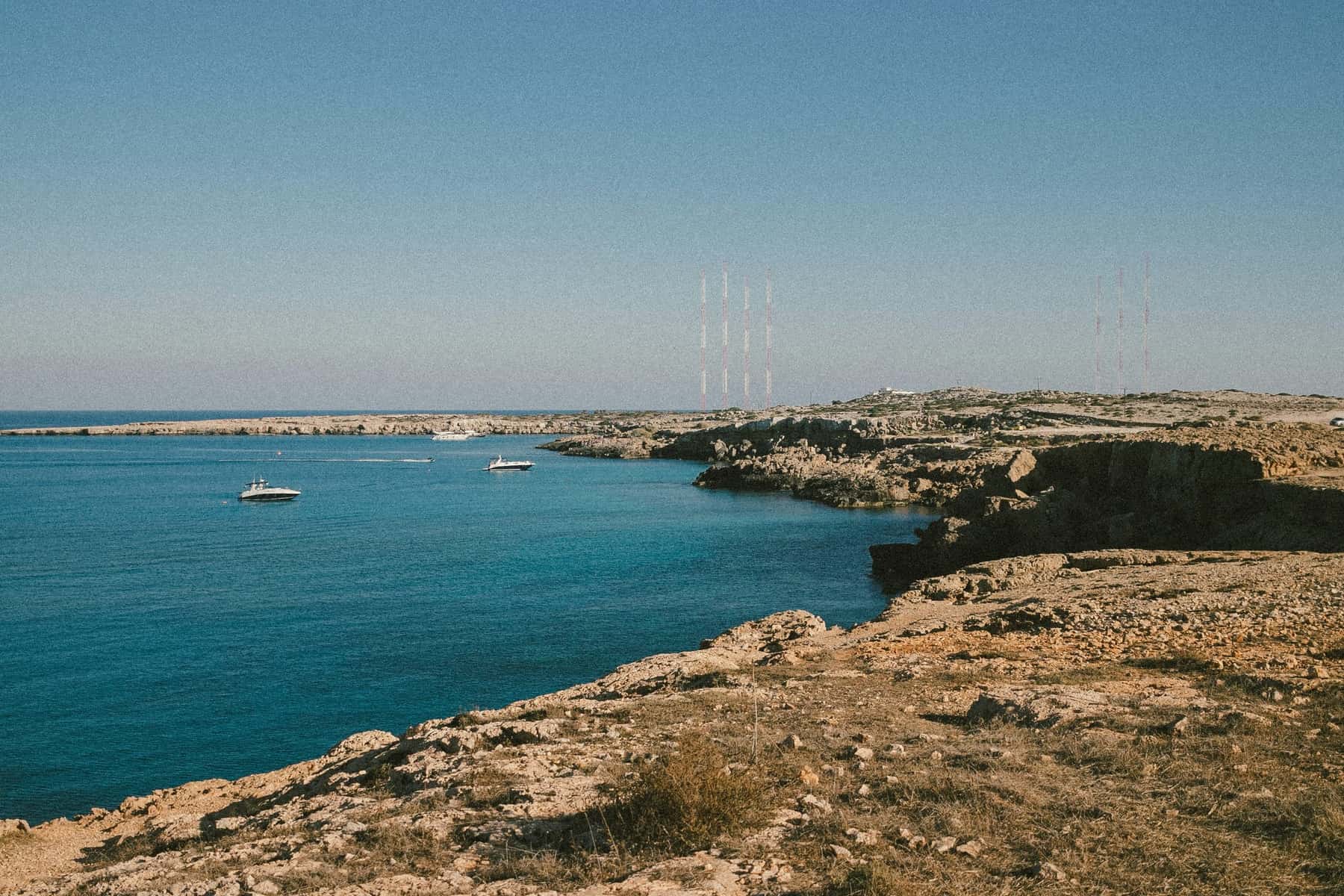 Rocky coastline beside a calm turquoise bay at Cape Greco, Cyprus, with small boats on the water
