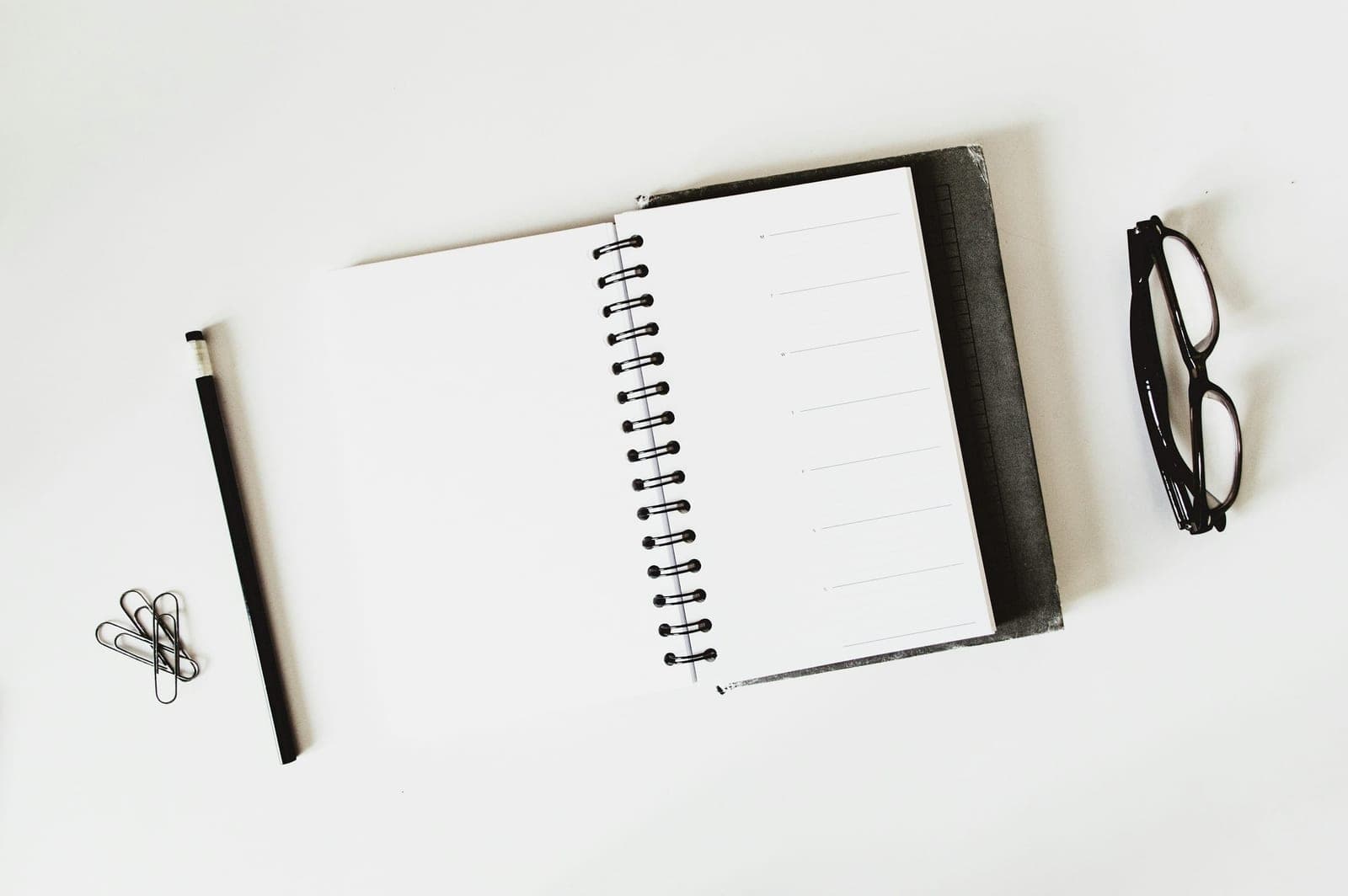 An open notebook, a pencil and a pair of glasses on a plain white desk